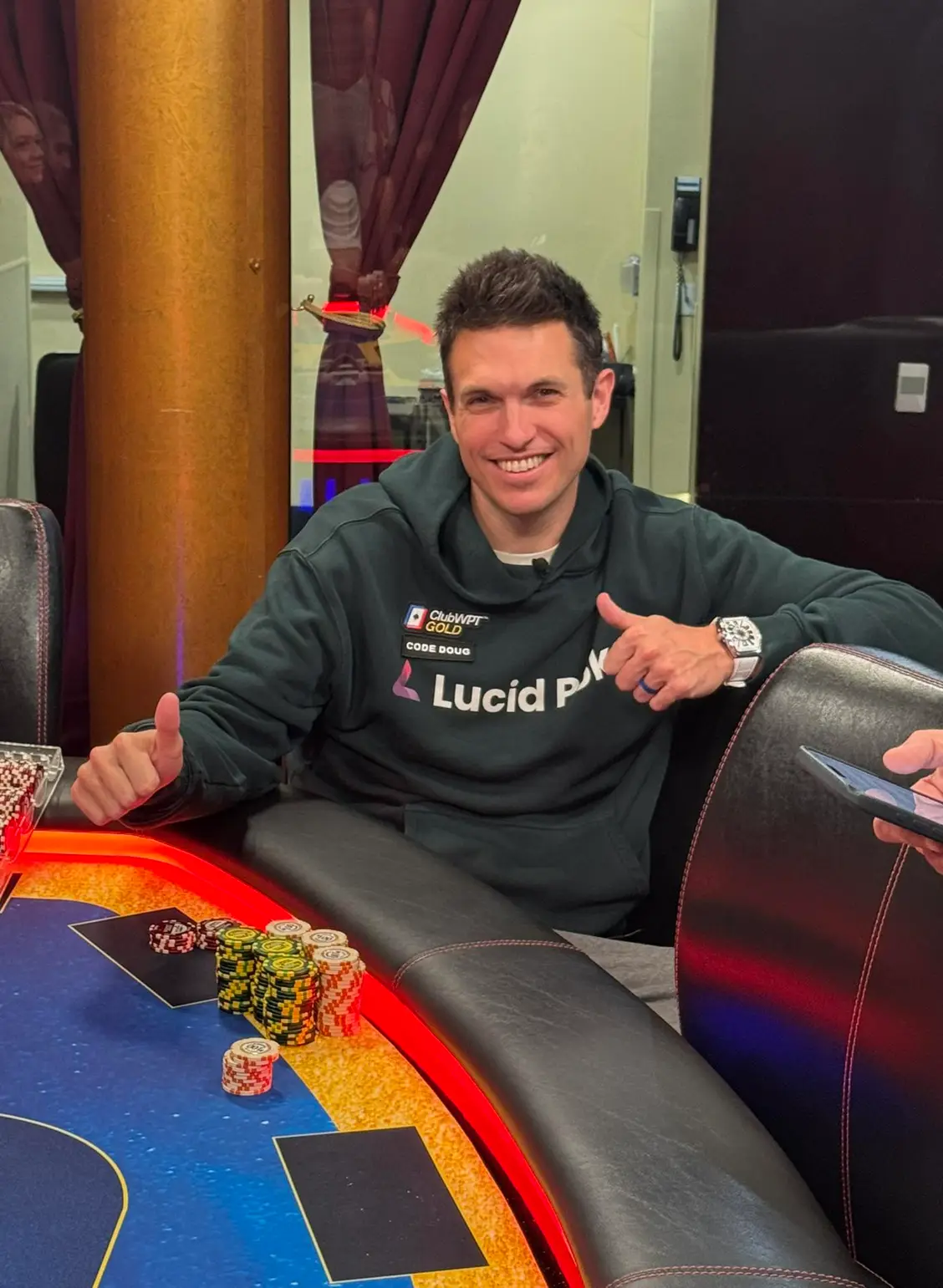 Doug Polk at a poker table wearing a ClubWPT Gold and Lucid Poker hoodie with chip stacks in front of him