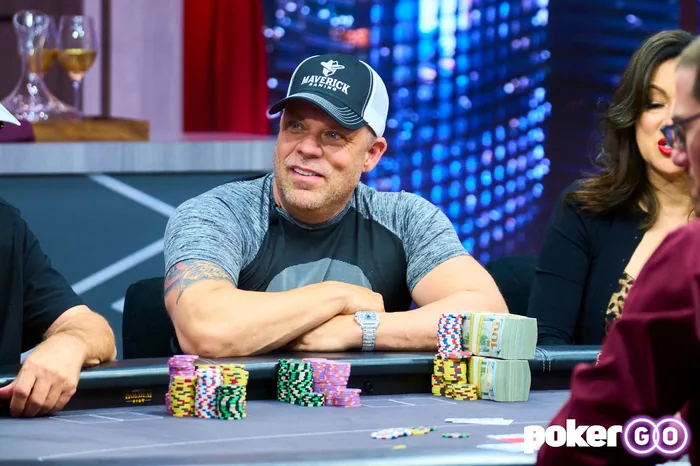Eric Persson sitting with arms crossed behind stacks of chips and cash bundles on a PokerGO broadcast