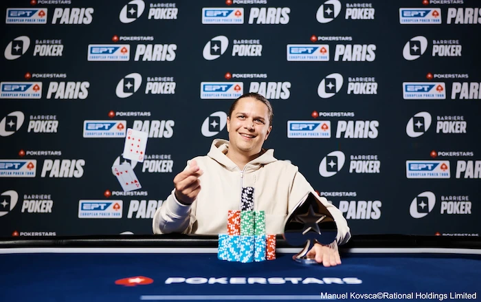 Niklas Astedt, Swedish professional poker player and high-roller specialist, smiling and holding up playing cards at the EPT Paris final table, wearing a light hoodie, with tall chip stacks in front against a branded PokerStars and EPT Paris backdrop.