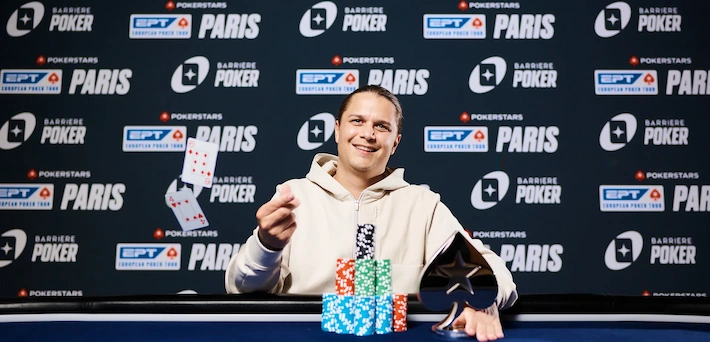 Niklas Astedt, Swedish professional poker player and high-roller specialist, smiling and holding up playing cards at the EPT Paris final table, wearing a light hoodie, with tall chip stacks in front against a branded PokerStars and EPT Paris backdrop.