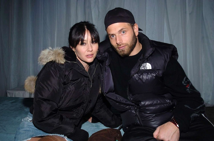 Rick Salomon and Shannen Doherty posing together at an event, him in a black The North Face puffer jacket and cap with beard, her in a black puffer jacket with fur hood and denim skirt, sitting close on a bed in a dimly lit room.