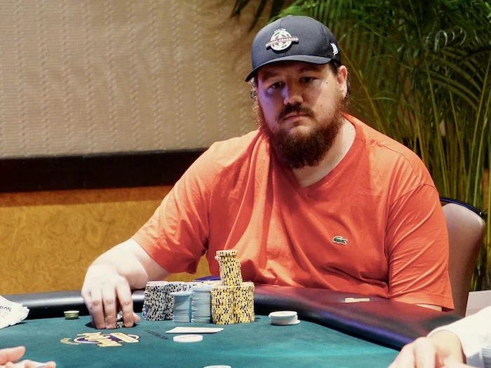 Shaun Deeb, elite mixed-game specialist, seated at the tournament table in orange Lacoste polo and navy cap, with tall chip stacks in front.