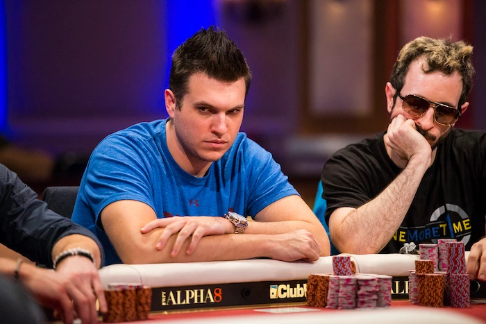 Doug Polk, 3x WSOP bracelet winner and high-stakes legend, intense at the poker table in blue t-shirt with spiky hair, arms crossed during live high-roller action.