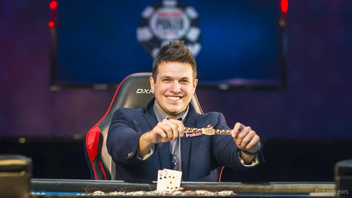 Doug Polk, 3x WSOP bracelet winner, smiling and holding up championship belt after a big win at the poker table in navy suit jacket and DXRacer chair