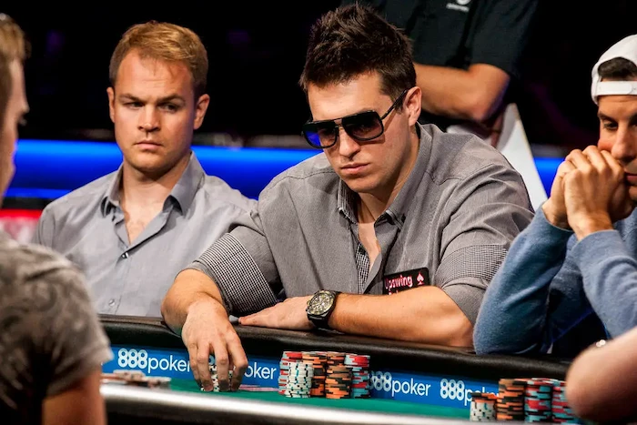Doug Polk, 3x WSOP bracelet winner and high-stakes cash game specialist, focused at the poker table in gray shirt and sunglasses, arms on rail with chip stacks during a televised high-roller session.