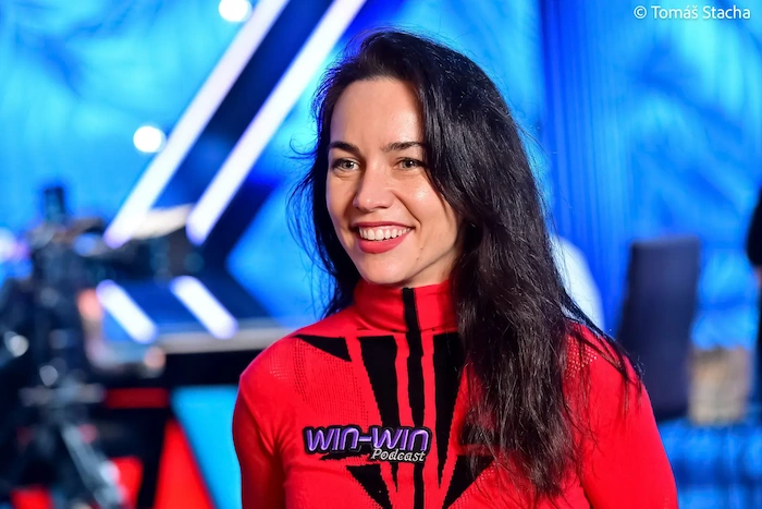 Liv Boeree, former professional poker player and EPT champion, smiling at the camera in a red WIN-WIN Podcast branded turtleneck top during a bright studio or event appearance