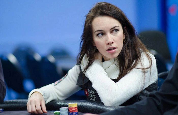 Liv Boeree, former professional poker player and EPT champion, leaning on the rail with hand on neck in white long-sleeve top and PokerStars vest, looking upward thoughtfully at the poker table during a live event.