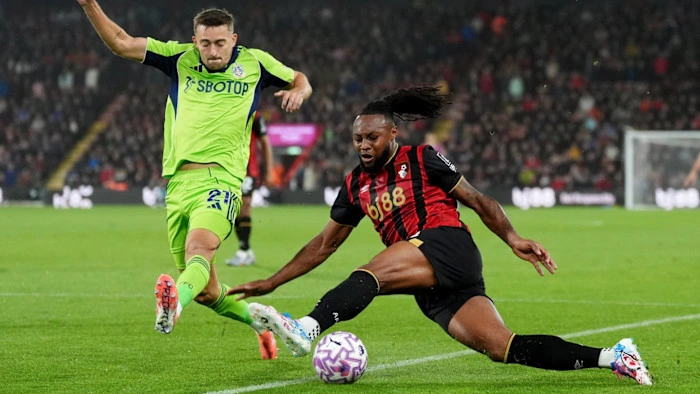 Bournemouth's bj88 Gambling Sponsor on Display Bournemouth player in red and black kit sliding to tackle, with hair flowing, as a Brentford player in neon green kit leaps over him to contest the ball on the pitch during a Premier League match under floodlights.