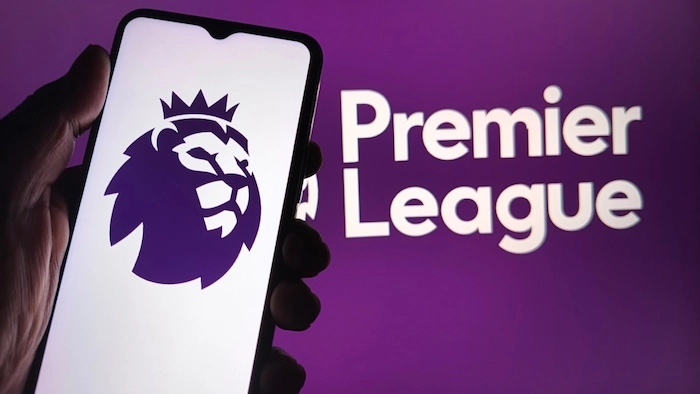 Smartphone screen displaying the Premier League logo (purple lion crown emblem) with "Premier League" text below, held in a hand against a matching purple background.