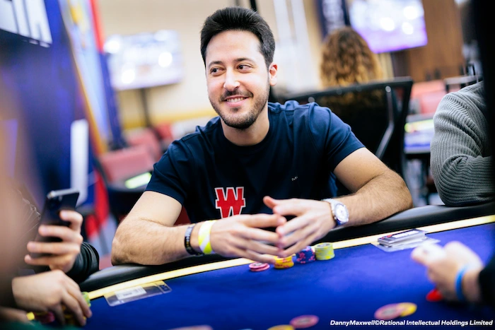 Adrian Mateos, Spanish poker pro and multiple WSOP bracelet winner, smiling at the WSOP table in a navy t-shirt with W logo, hands clasped on the rail with colorful chip stacks, surrounded by blurred players and blue/purple event lighting.