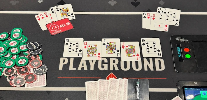 Playground poker table showdown with K-high straight flush vs J-high straight flush, all-in marker, scattered hole cards, colorful chip stacks in green/red/black, electronic dealer button and shuffler on black felt mat.