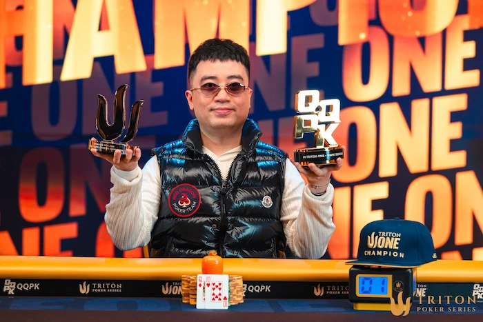 Nguyen Huu Dung, Vietnamese poker pro, holding up two Triton Poker trophies (one horn-shaped, one with "TRITON CHAMPION" plaque) at the Triton Series final table, wearing tinted sunglasses, blue-black puffer vest over white shirt with patches, smiling against an orange-blue "TRITON POKER SERIES CHAMPION" backdrop.